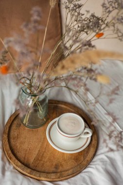 Hot mug of cappuccino on wooden tray on the bed, breakfast, bouquet of dried flowers. Spring scenery. Cozy house.