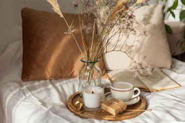 Cozy breakfast in bed. Cup of hot cappuccino on wooden tray, bouquet of dried flowers in glass vase, book, eco gift box, candle.