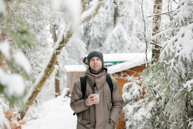 Portrait of young man in warm clothes in winter forest. Drinking hot tea outdoors from thermos. Hiking.