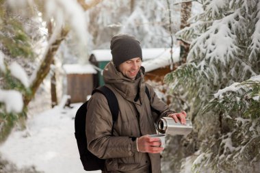 Portrait of young man in warm clothes in winter forest. Drinking hot tea outdoors from thermos. Hiking.