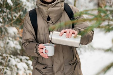 Portrait of young man in warm clothes in winter forest. Drinking hot tea outdoors from thermos. Hiking.