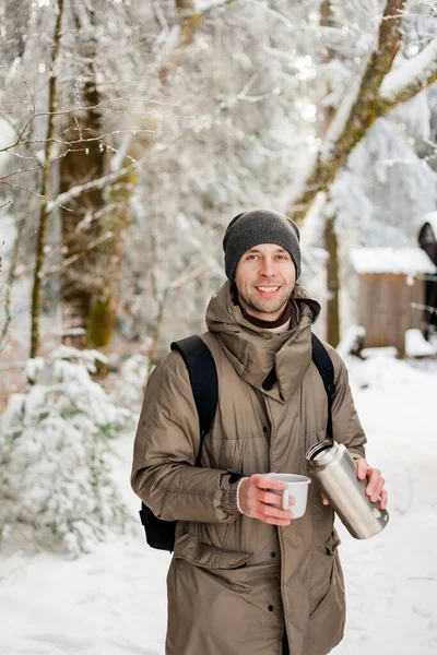 Portrait of young man in warm clothes in winter forest. Drinking hot tea outdoors from thermos. Hiking.