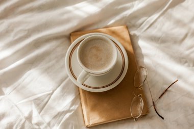 A cup of hot cappuccino, a book, glasses on the bed. Breakfast. Cozy house. Decor.