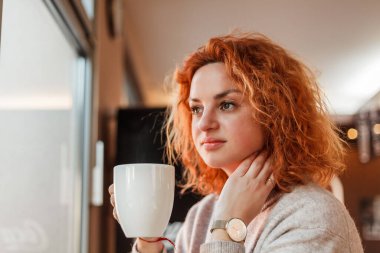 Red-haired curly young bright woman with green eyes drinks hot coffee in cozy cafe. Beautiful model in stylish clothes.