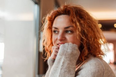 Red-haired curly young bright woman with green eyes drinks hot coffee in cozy cafe. Beautiful model in stylish clothes.