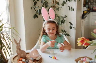 Easter. A cute little girl in bunny ears decorates Easter eggs at the table at home. Spring.