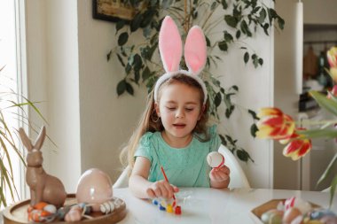Easter. A cute little girl in bunny ears decorates Easter eggs at the table at home. Spring.