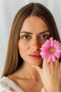 Beyaz arka planda pembe gerberalı güzel, uzun saçlı, kahverengi gözlü bir kadının portresi. Bahar. Tatiller.