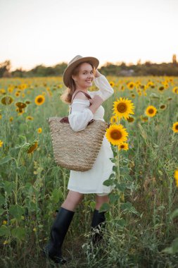 Portrait of beautiful young woman 33 years old in hat in sunflower field at sunset. Happy model in white dress on summer evening in nature.