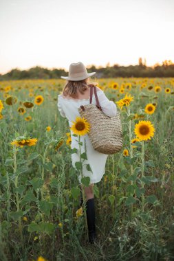 Portrait of beautiful young woman 33 years old in hat in sunflower field at sunset. Happy model in white dress on summer evening in nature.
