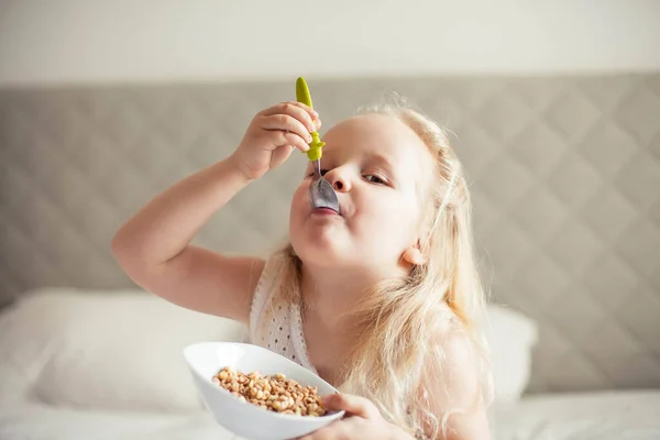 小さなかわいい白い髪の女の子は彼女の明るい部屋のベッドの上にグラノーラを食べる 健康食品 — ストック写真