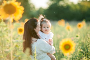 Koyu renk saçlı güzel anne, 1,5 yaşındaki kızıyla güneşin altındaki ayçiçeği tarlasında oynuyor. Aile.