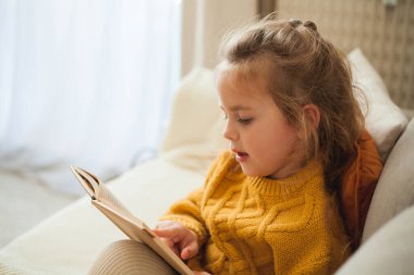 Örgü turuncu süveter giyen 4 yaşındaki tatlı küçük kız rahat bir odada kitap okuyor. Düş.
