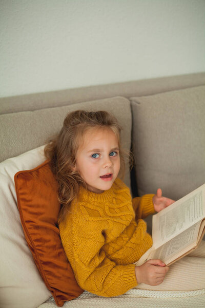 Cute little girl of 4 years old in knitted orange sweater is reading a book in a cozy room. Fall.