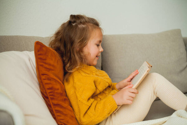Cute little girl of 4 years old in knitted orange sweater is reading a book in a cozy room. Fall.