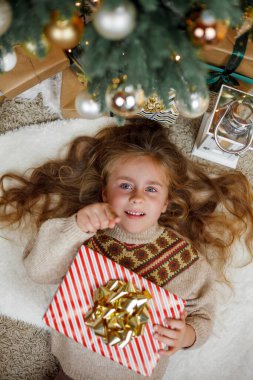 5 yaşında, uzun kıvırcık saçlı ve mavi gözlü sevimli bir kız Noel ağacının yanındaki hediye kutularını açıyor. Tatillerde. Yeni yıl. Kış tatili.