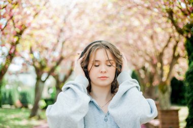 Beautiful young woman in comfortable clothes listens to music in headphones in blooming park. Sakura Pink flowers.