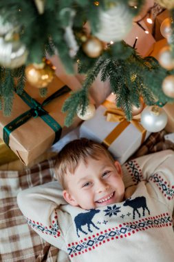 Happy boy open gifts near the Christmas tree. clipart