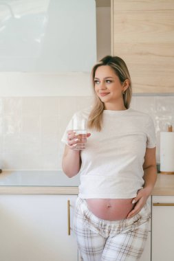 Pijamalı hamile bir kadın ellerinde bir bardak su tutuyor ve mutfakta onu içiyor. Sağlıklı vücut.
