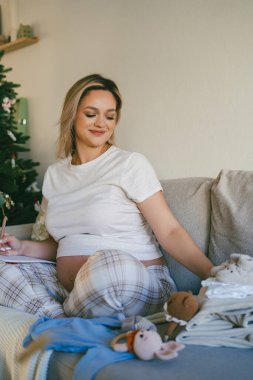 Pijamalı güzel bir kadın doğum koğuşu için bebek eşyalarıyla dolu bir çanta hazırlıyor. Göbekli manken anne olmaya hazırlanıyor..