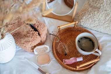 Hasır tablada bir fincan sıcak kahve, Boucle kozmetik poşeti, beyaz çarşaflarda kozmetik ürünler. Bahar havası. Ev ışığı bej iç dekorasyon.