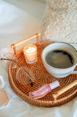 Hasır tablada bir fincan sıcak kahve, Boucle kozmetik poşeti, beyaz çarşaflarda kozmetik ürünler. Bahar havası. Ev ışığı bej iç dekorasyon.