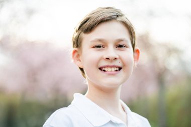 10 yaşında, Avrupa parkında beyaz polo giyen, pembe sakura çiçekli sevimli kahverengi gözlü bir çocuğun portresi. Bahar havası.