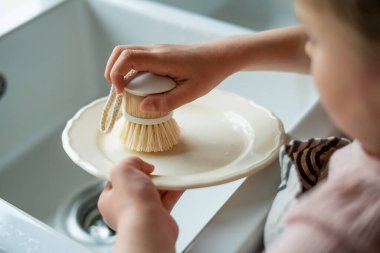 Mutfaktaki lavabonun yanında bir tabak yıkayan çocukların elleri. Küçük kız elinde bir bulaşık fırçası tutuyor..