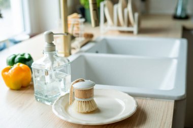 Lavabonun yanında bulaşık yıkamak için bir ekofırça, cam şişede deterjan ve havlu. Parlak mutfak iç mimarisi.