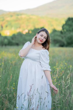 Hamile genç bir kadın, bebek beklentisiyle ellerini karnının üstüne koyup sahada yürüyor. Mutlu annelik.