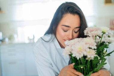Koyu saçlı genç bir kadın parlak bir mutfaktaki vazoya pembe bir buket çiçek koyar. Samimi.