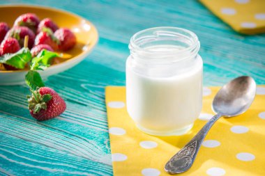Turkuaz renkli ahşap masada bir kavanoz yoğurt ve çilek.