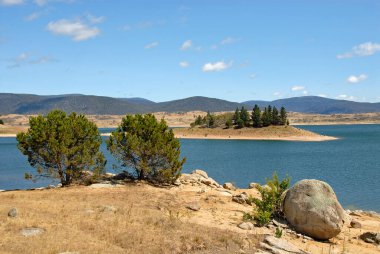 Jindabyne, New South Wales, Avustralya: Önünde ağaçlar ve kayalar olan Jindabyne Gölü 'ne bakın. Jindabyne, Karlı Dağlar yakınlarında bir turizm merkezidir..