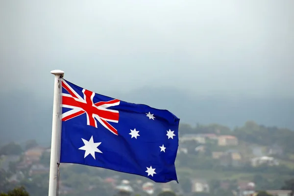 Australian flag set against a background of houses on a hill and sky. The Australian flag features six stars and the British union jack.