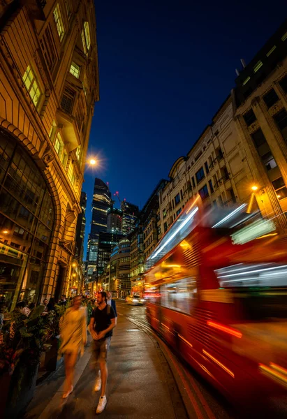 stock image London, UK - June 14 2023: Gracechurch Street in the City of London at night. People walk along the pavement as a red London bus passes by. Tall buildings in the distance.