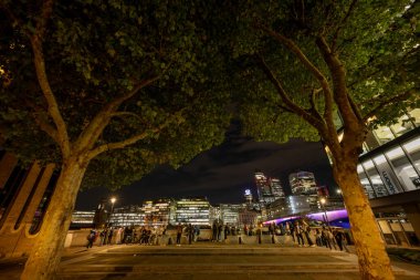 Londra, İngiltere: Londra şehrinin gece manzarası Thames Nehri 'nin güney kıyısından Southwark' taki Londra Köprüsü 'ne doğru çekildi. Ön plandaki insanlar ağaçlarla çevrili..