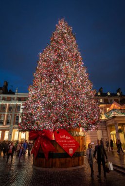 Londra, İngiltere - 8 Kasım 2023: Covent Garden Market 'in dışındaki büyük süslü Noel ağacı. İnsanlar alışveriş poşetleriyle yanımızdan geçiyor. Portre yönelimi.