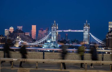 Londra, İngiltere: Londra Köprüsü 'nde geceleri hareket bulanıklığı ile yürüyen insanlar. Londra rıhtımının arkasındaki Kule Köprüsü ve gökdelenler.
