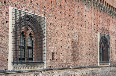 İtalya 'nın Milano kentindeki tarihi Sforza Şatosu' nun (Castello Sforzesco) ağır demir ızgarası tarafından korunan ortaçağ tuğlalı bir duvardaki Gotik tarzı kemerli pencerenin yakın çekimi,
