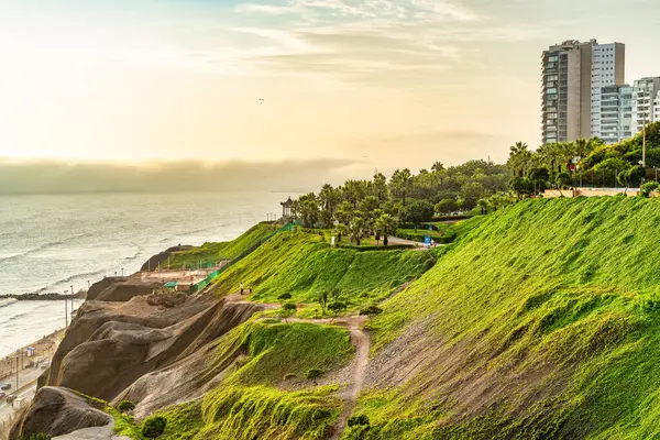 Peru 'nun Lima bölgesindeki Miraflores bölgesinde yemyeşil bir uçurum parkının manzarası. İnsanlar gün batımında sahil yolunda yürüyorlar..