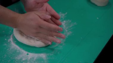 Düz un hamurunu şekillendiren ve pizza yapmaya hazırlayan bir çift el. Sahne, hamuru esnetme ve şekillendirme sürecini vurguluyor. Üniformalı bir pizza tabanının malzemelerle doldurulmaya hazır olması için gereken beceriyi gösteriyor..