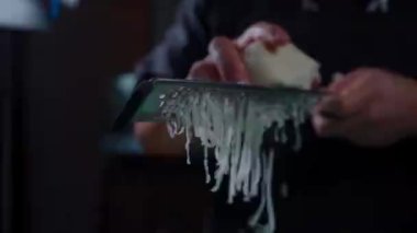 Peynir rendesinin yakın çekimini yaparken taze mozzarella ekleyip rendelenmiş peynirin dokusunu ve akışını vurguluyor. Görüntü, hazırlıktaki hassasiyeti ve malzemenin kalitesini gösteriyor.