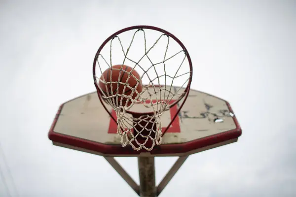 Düşük açılı bir perspektiften çekilen basketbol sepetinin detaylı görüntüsü ve arka tahtası kötü durumda. Bu görünüm, hem kırmızı hem de tahtanın uzun süreli kullanımının etkisini gösteren aşınma ve bozulmayı vurgular..