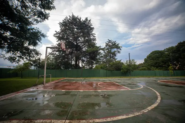 cancha de baloncesto mojada despus de la lluvia, con el agua acumulada en el suelo reflejando el entorno. El fondo combina elementos naturales y urbanos, capturando el contraste entre la cancha deportiva y el paisaje circundante.
