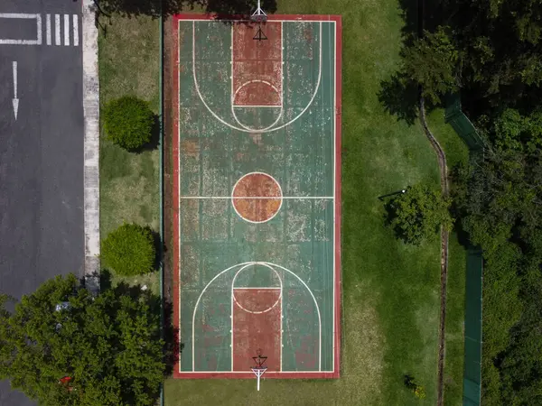 Doğanın kentsel manzarayla karıştığı bir ortamda bulunan bir basketbol sahasının havadan çekimi. Mahkemenin doğal ve kentsel unsurları birleştirerek benzersiz bir görsel karşıtlık yaratan bir alanda bulunduğu gözlemlenmektedir.
