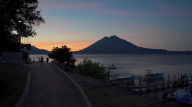 Atitlan Gölü 'nde gün doğumu. Birkaç tekne, ahşap bir rıhtımın yanında huzur içinde dinleniyor. Güneşin ilk ışınları ufku aydınlatır ve gölün sakin sularına yansıtır.