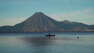 Teknesinde bir adam, Atitlan Gölü 'nün sakin şafağında balık tutmaya hazırlanıyor. Yeni doğan güneşin yumuşak ışığı, ufuktaki volkanlar görkemli bir şekilde yükseldikçe suyu aydınlatır..
