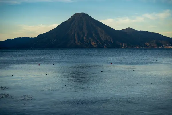 Atitlan Gölü kıyılarında baş döndürücü bir gün doğumu. Güneşin ilk ışınları manzarayı nazikçe aydınlatıyor. Arka planda, volkan görkemli bir şekilde duruyor, doğal güzelliklerle dolu huzurlu bir manzara yaratıyor..