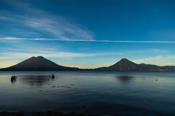 Atitlan Gölü kıyılarında, gölün etrafını saran dağların olduğu büyüleyici bir gün batımı manzarası. Batan güneşin sıcak renkleri suya yansıyarak barış ve doğal güzellik atmosferi yaratıyor.