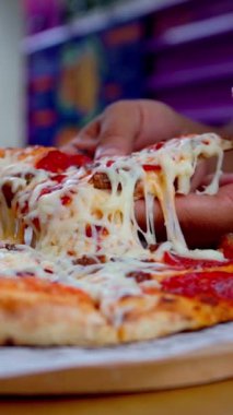 Sıcak bir dilim pizzayı kaldırıp erimiş peynirin mükemmelliğe nasıl uzandığını gösteren bir el. Altın hamuru ve taze malzemeler her ısırıktaki cazibeyi ortaya çıkarır..